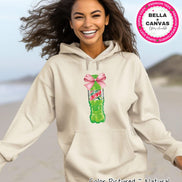 A natural-colored hoodie featuring a graphic of a Mountain Dew bottle with a pink bow. The hoodie has a front pocket and a drawstring hood. The image also includes the text 