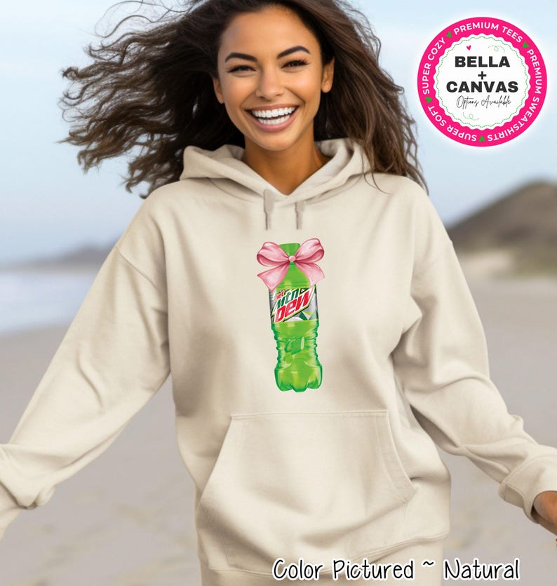 A natural-colored hoodie featuring a graphic of a Mountain Dew bottle with a pink bow. The hoodie has a front pocket and a drawstring hood. The image also includes the text 