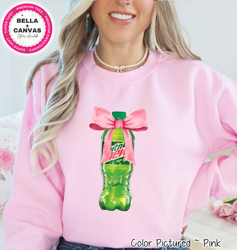 Pink sweatshirt featuring a graphic of a green soda bottle with a pink bow. The bottle has a green cap and the words "Mtn Dew" on it. The text "Color Pictured ~ Pink" is at the bottom. The sweatshirt has the Bella + Canvas logo.