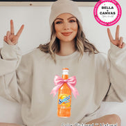 A beige sweatshirt featuring a watercolor illustration of a Sunkist Orange soda bottle tied with a pink bow. The bottle is labeled 
