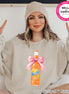 A beige sweatshirt featuring a watercolor illustration of a Sunkist Orange soda bottle tied with a pink bow. The bottle is labeled "Sunkist Orange 12 OZ". The text "Color Pictured ~ Natural" is at the bottom.