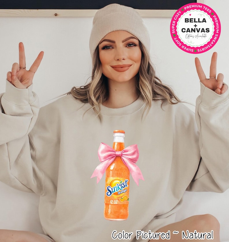 A beige sweatshirt featuring a watercolor illustration of a Sunkist Orange soda bottle tied with a pink bow. The bottle is labeled "Sunkist Orange 12 OZ". The text "Color Pictured ~ Natural" is at the bottom.
