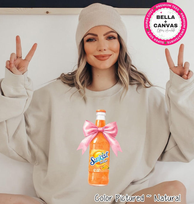 A beige sweatshirt featuring a watercolor illustration of a Sunkist Orange soda bottle tied with a pink bow. The bottle is labeled "Sunkist Orange 12 OZ". The text "Color Pictured ~ Natural" is at the bottom.