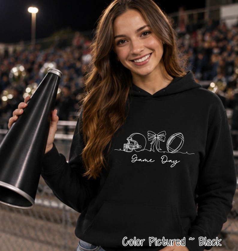 May include: A black hooded sweatshirt featuring a white line art graphic of a football helmet, a bow, and a football, with the text Game Day written below the design. The hoodie includes a front kangaroo pocket and long sleeves.