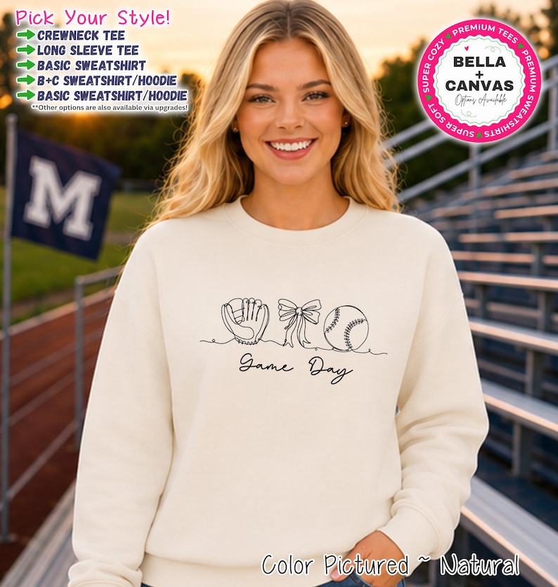 May include: A natural-colored crewneck sweatshirt featuring a black line art graphic of a baseball glove, a bow, and a baseball, with the text 