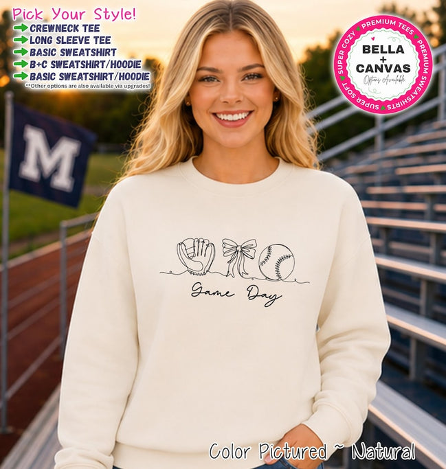 May include: A natural-colored crewneck sweatshirt featuring a black line art graphic of a baseball glove, a bow, and a baseball, with the text "Game Day" written below the icons. The image includes text options for style and brand information.