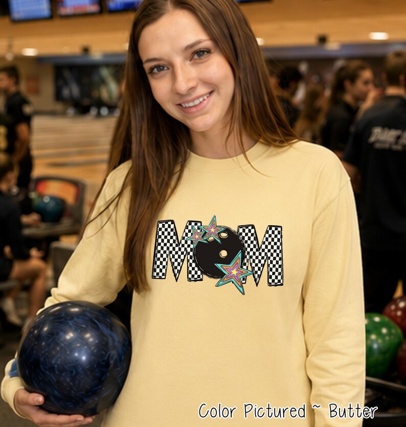 May include: A long-sleeve butter yellow shirt featuring a graphic print of the word MOM in a black and white checkered pattern. The letter O is replaced by a black bowling ball, and the design is accented with colorful star illustrations.