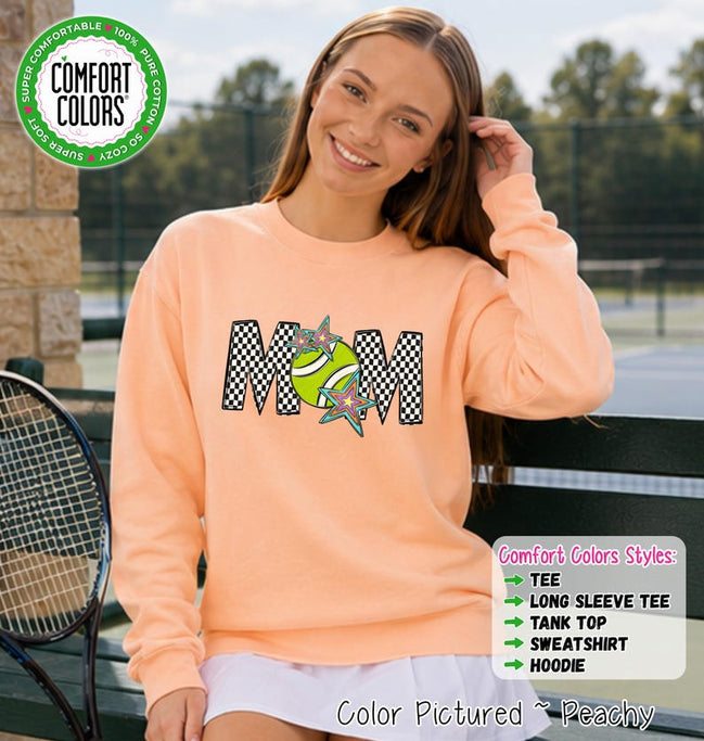 May include: A peach-colored long-sleeve sweatshirt featuring the text MOM in a black and white checkered pattern. The letter O is replaced by a lime green tennis ball graphic adorned with two colorful, multi-pointed star shapes.