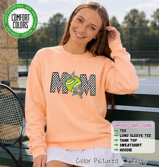 May include: A peach-colored long-sleeve sweatshirt featuring the text MOM in a black and white checkered pattern. The letter O is replaced by a lime green tennis ball graphic adorned with two colorful, multi-pointed star shapes.