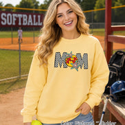May include: A butter yellow crewneck sweatshirt featuring the word MOM in a black and white checkered pattern. The letter O is replaced by a yellow softball graphic with red stitching, accented by two colorful, multi-pointed star shapes.
