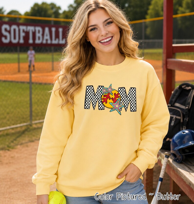 May include: A butter yellow crewneck sweatshirt featuring the word MOM in a black and white checkered pattern. The letter O is replaced by a yellow softball graphic with red stitching, accented by two colorful, multi-pointed star shapes.
