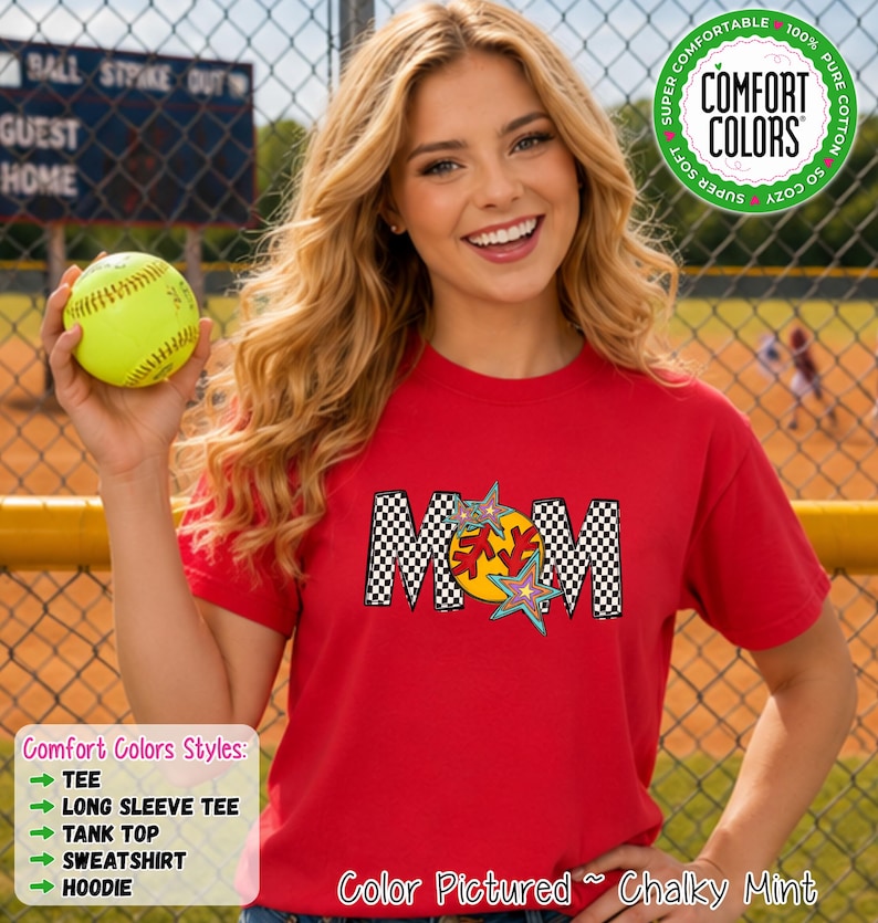 May include: A red short-sleeve t-shirt featuring a graphic with the word MOM in a black and white checkered pattern. The letter O is replaced by a yellow softball with red stitching, accented by two colorful star shapes. A green circular badge reads COMFORT COLORS.