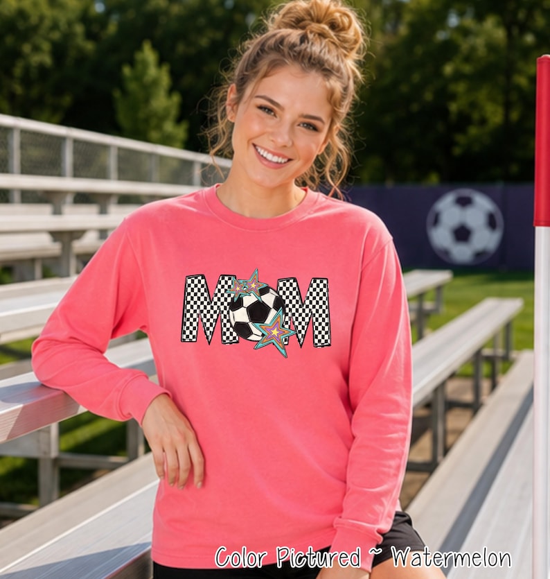 May include: A watermelon-colored long-sleeve sweatshirt featuring the word MOM in a black and white checkered pattern. The letter O is replaced by a black and white soccer ball graphic, accented with two colorful, multi-pointed star shapes.