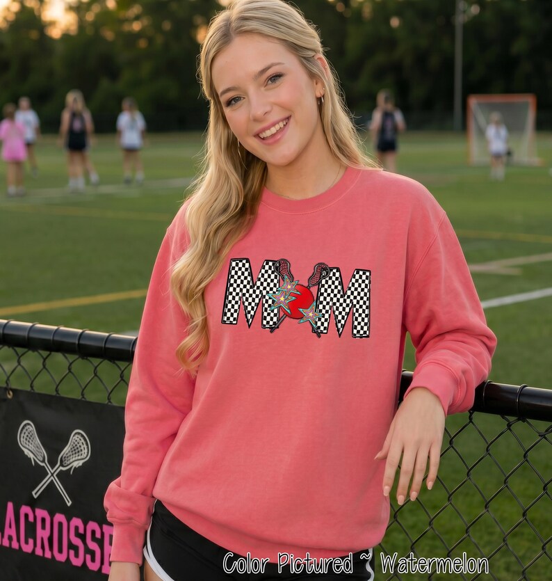 May include: A watermelon-colored crewneck sweatshirt featuring the word MOM in a black and white checkered pattern. The letter O is replaced by a red lacrosse ball with two crossed lacrosse sticks behind it, accented by two small blue star shapes.