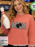 May include: A terracotta-colored crewneck sweatshirt featuring a graphic print of the word MOM in a black and white checkerboard pattern, with a black bowling ball replacing the letter O and colorful star accents surrounding the text.