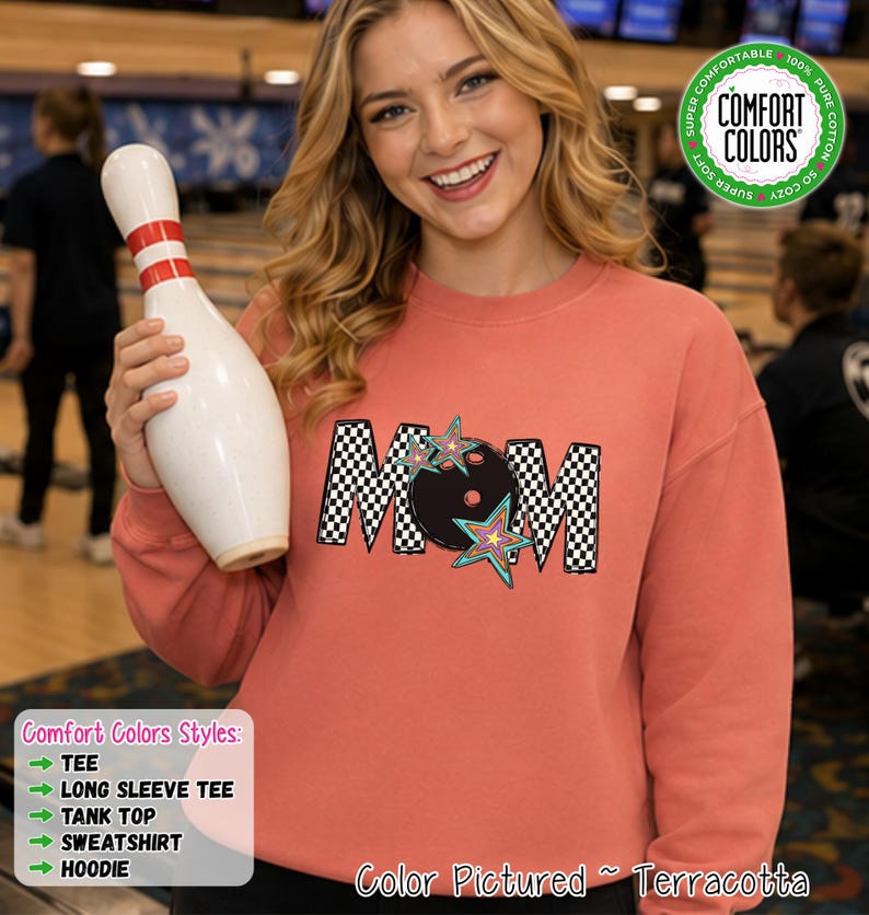 May include: A terracotta-colored crewneck sweatshirt featuring a graphic print of the word MOM in a black and white checkerboard pattern, with a black bowling ball replacing the letter O and colorful star accents surrounding the text.