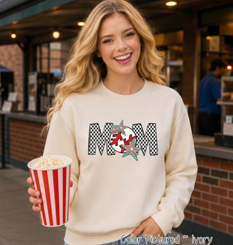 May include: An ivory crewneck sweatshirt featuring a graphic print of the word MOM in a black and white checkered pattern. The letter O is replaced by a red and white baseball design, accented with two colorful, multi-pointed star shapes.