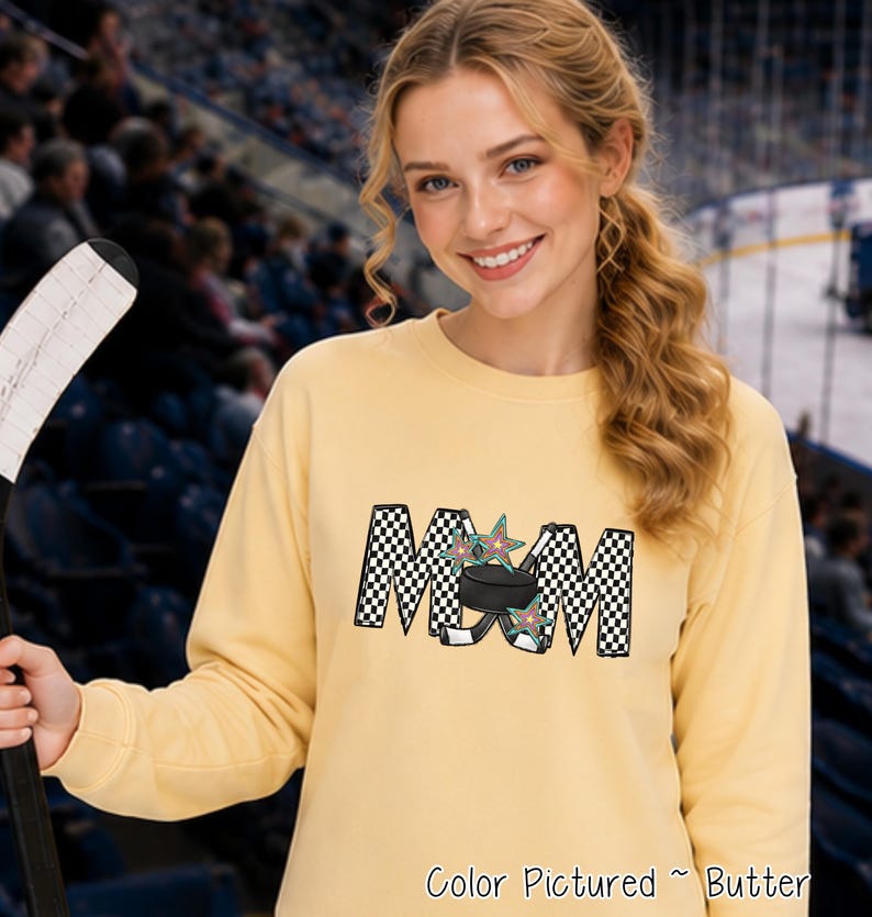 May include: A butter yellow long-sleeve sweatshirt featuring a graphic print of the word MOM in a black and white checkered pattern. The letter O is replaced by a black hockey puck and two crossed hockey sticks with colorful star accents.