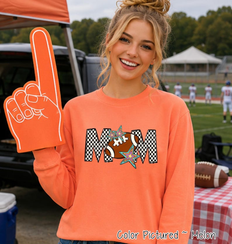 May include: A melon-colored long-sleeve sweatshirt featuring a graphic print of the word MOM in a black and white checkered pattern. The letter O is replaced by a brown football with white laces, accented by two colorful star shapes.