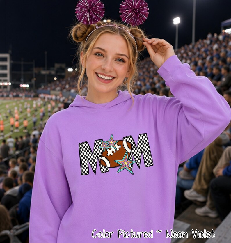 May include: A neon violet hooded sweatshirt featuring a graphic print of the word MOM in a black and white checkered pattern. The letter O is replaced by a brown football, and the design is accented with two colorful, multi-pointed stars.