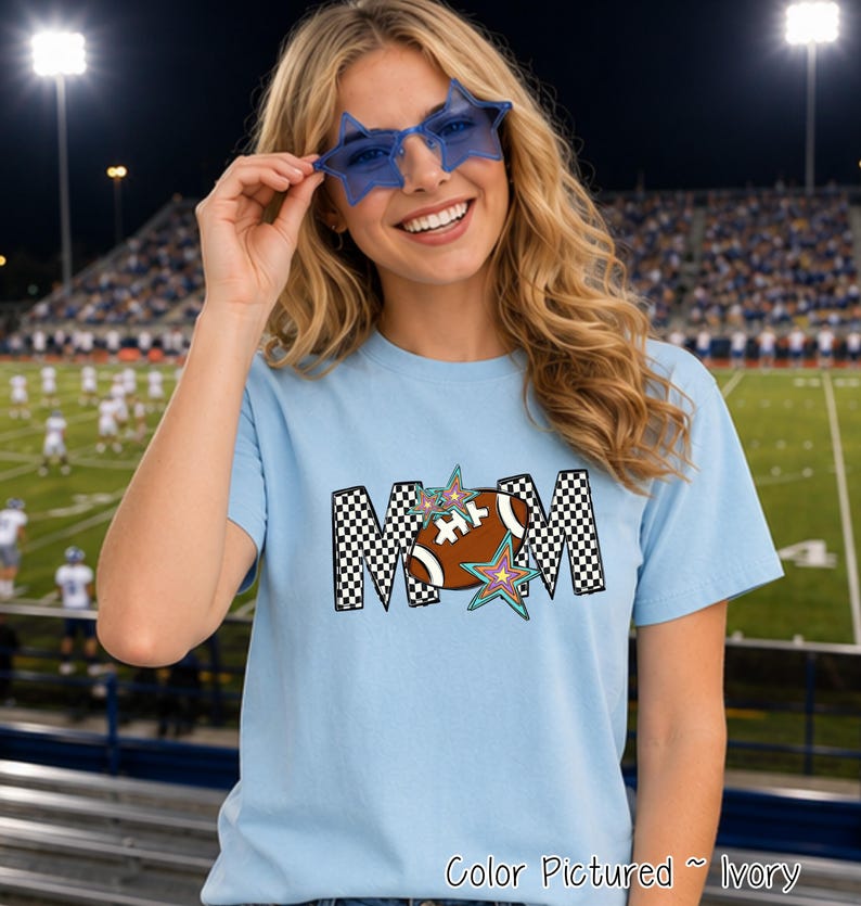 May include: A light blue short-sleeved t-shirt featuring a graphic print of the word MOM in a black and white checkered pattern. The letter O is replaced by a brown football with white laces, accented by two colorful star illustrations.