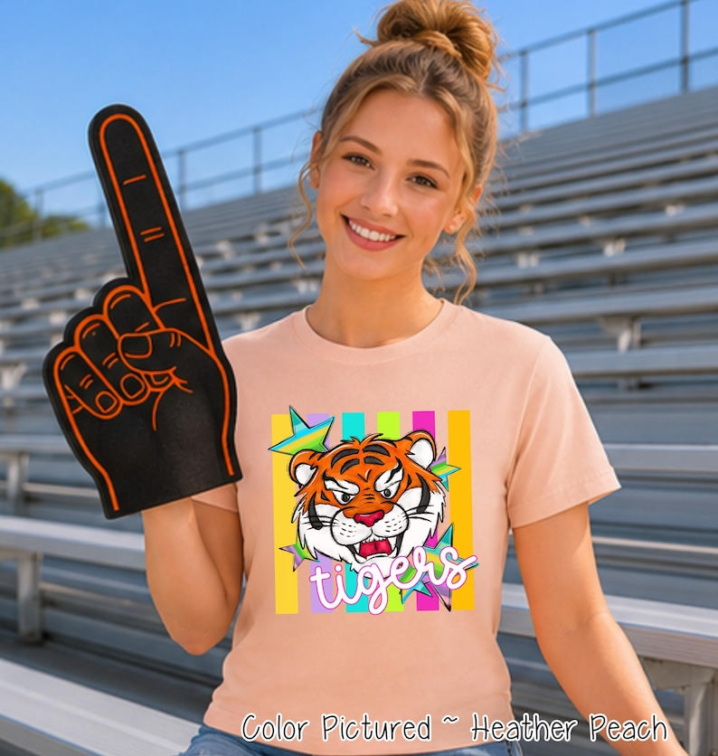May include: A heather peach t-shirt featuring a graphic of a tiger face surrounded by colorful stars and vertical stripes. The word "tigers" is printed in a white script font below the tiger. A black foam finger with orange trim is also shown.