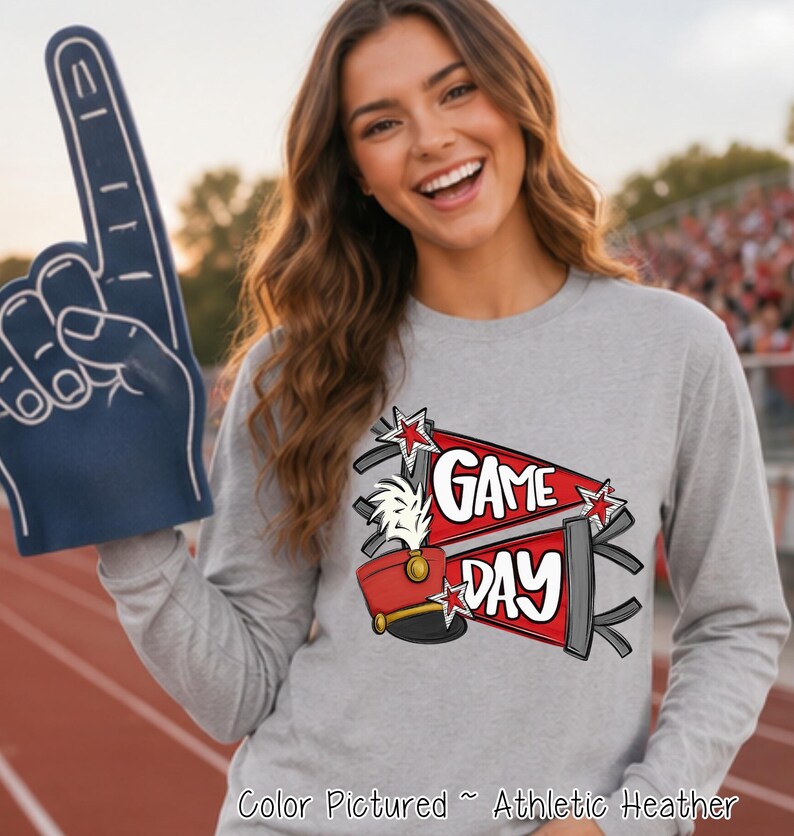 May include: An athletic heather gray long-sleeve shirt featuring a graphic print of two red pennants with the text "GAME DAY" in white, accompanied by a red marching band hat with a white plume and small star accents, held with a blue foam finger.