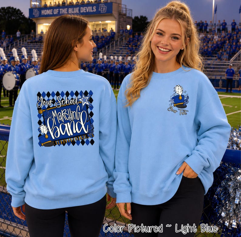 May include: A light blue crewneck sweatshirt featuring a marching band graphic. The back displays a blue and black diamond pattern with the text Your School Marching Band. The front left chest shows a blue marching band hat with a white plume.