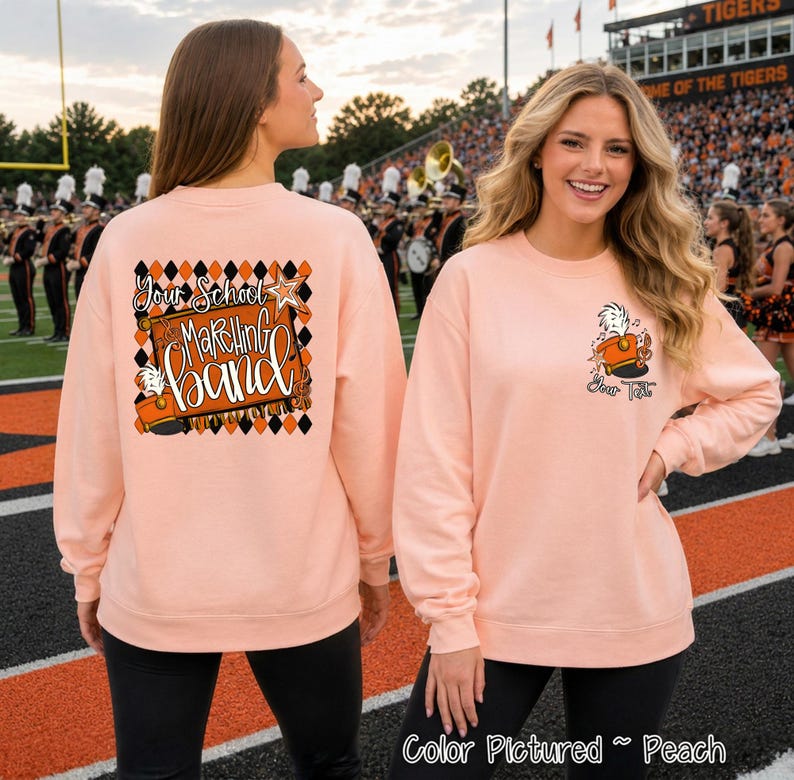 May include: A peach-colored crewneck sweatshirt featuring a large graphic on the back with a black and orange diamond pattern, a marching band hat, and the text 