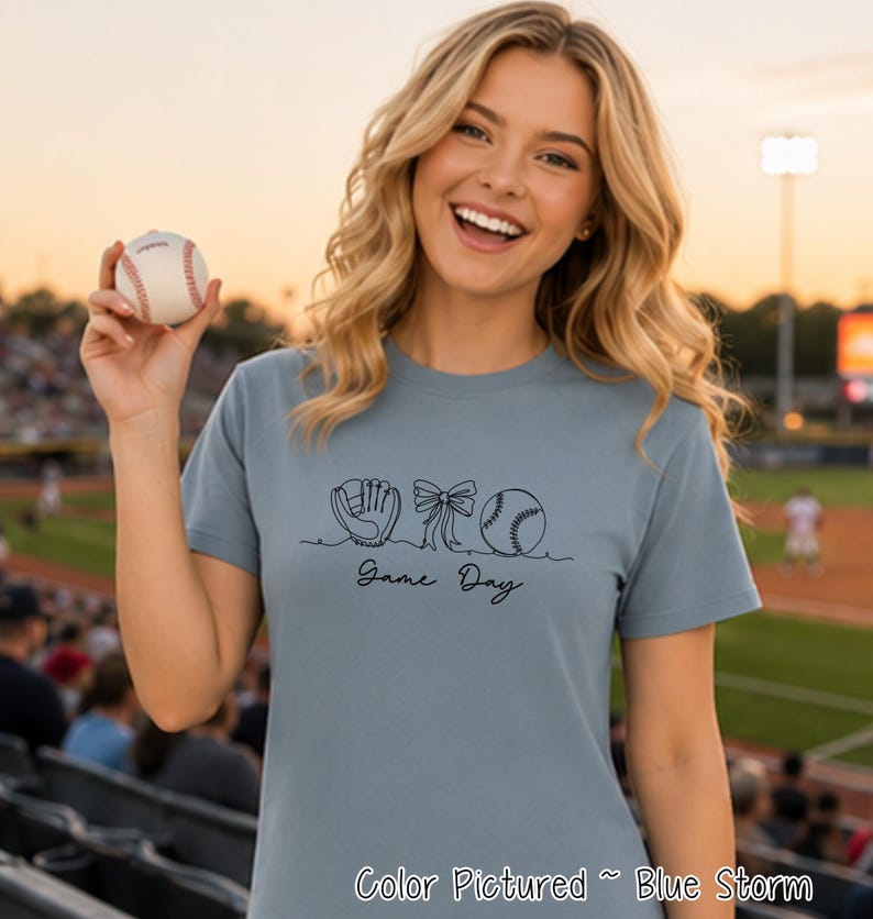 May include: A blue storm colored short-sleeve t-shirt featuring a black line art graphic of a baseball glove, a bow, and a baseball, with the text "Game Day" written in a cursive script font below the illustrations.