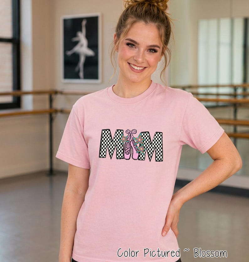 May include: A light pink short-sleeve t-shirt featuring the text MOM in a black and white checkered pattern. The letter O is replaced by a pair of pink ballet slippers with teal star accents and a pink bow. The shirt is a solid light pink color.