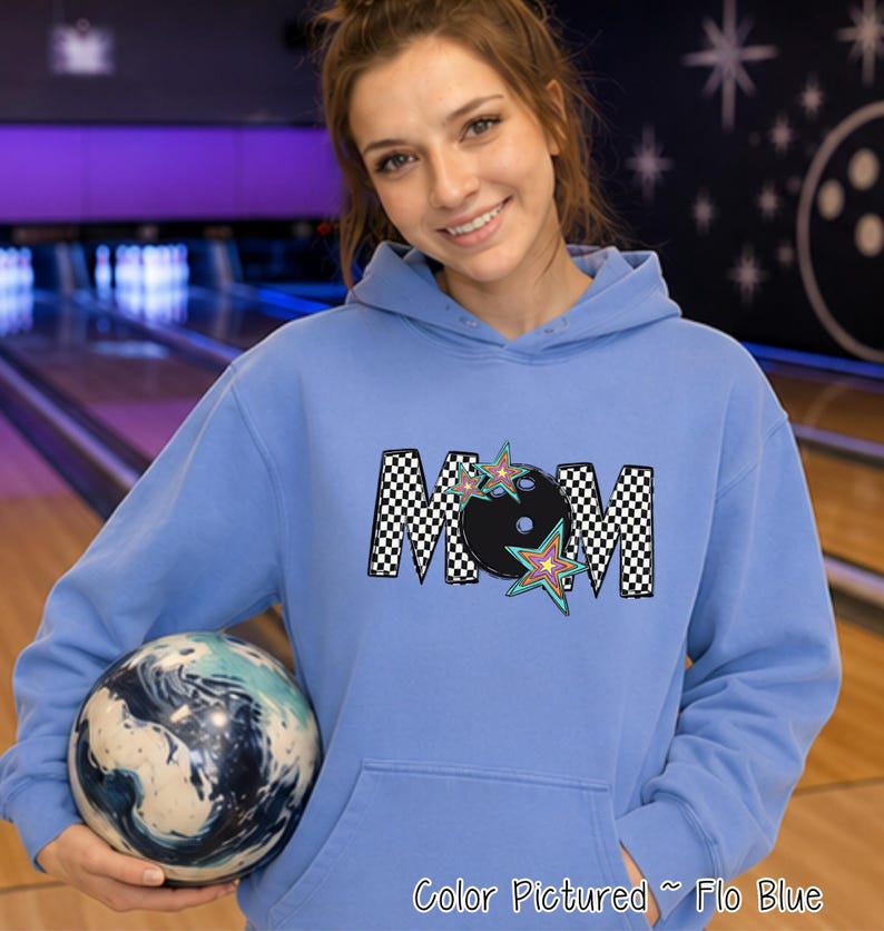 May include: A light blue hooded sweatshirt featuring a graphic print on the chest. The design displays the word MOM in a black and white checkered pattern, with a black bowling ball replacing the letter O and colorful star accents.