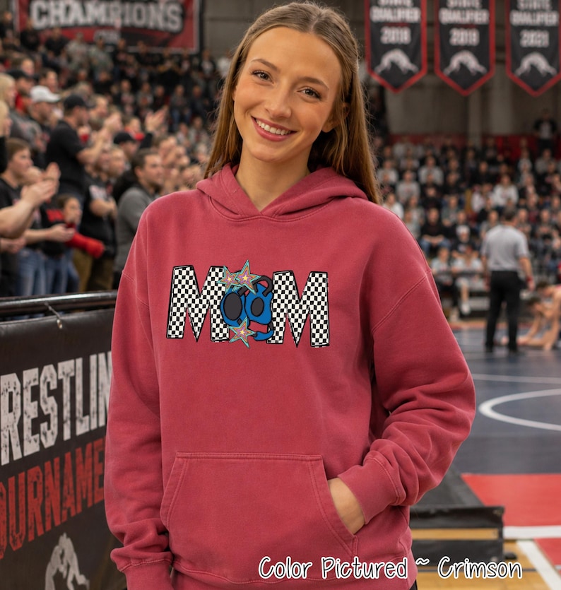 May include: A crimson hooded sweatshirt featuring a graphic on the chest that spells MOM in a black and white checkered pattern. The letter O is replaced by a blue wrestling headgear design, and two multi-colored stars are placed above and below it.
