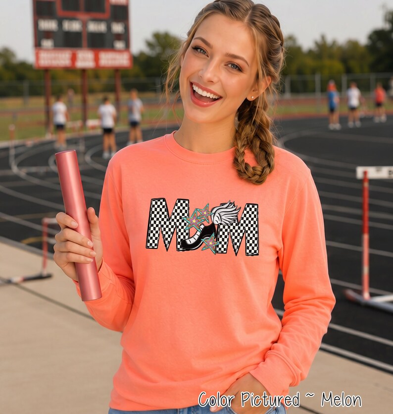 May include: A long-sleeve melon-colored sweatshirt featuring the word MOM in a black and white checkered pattern. The letter O is replaced by a black track shoe with a white wing and teal star accents. The sweatshirt is held with a pink relay baton.