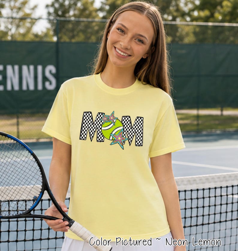 May include: A neon lemon yellow short-sleeve t-shirt featuring the word MOM in a black and white checkered pattern. The letter O is replaced by a green tennis ball graphic, and two colorful star shapes are layered over the tennis ball design.