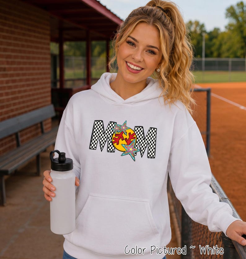 May include: A white hooded sweatshirt featuring a graphic print on the chest. The design displays the word MOM in a black and white checkered pattern, with a yellow softball replacing the letter O and colorful star accents surrounding it.