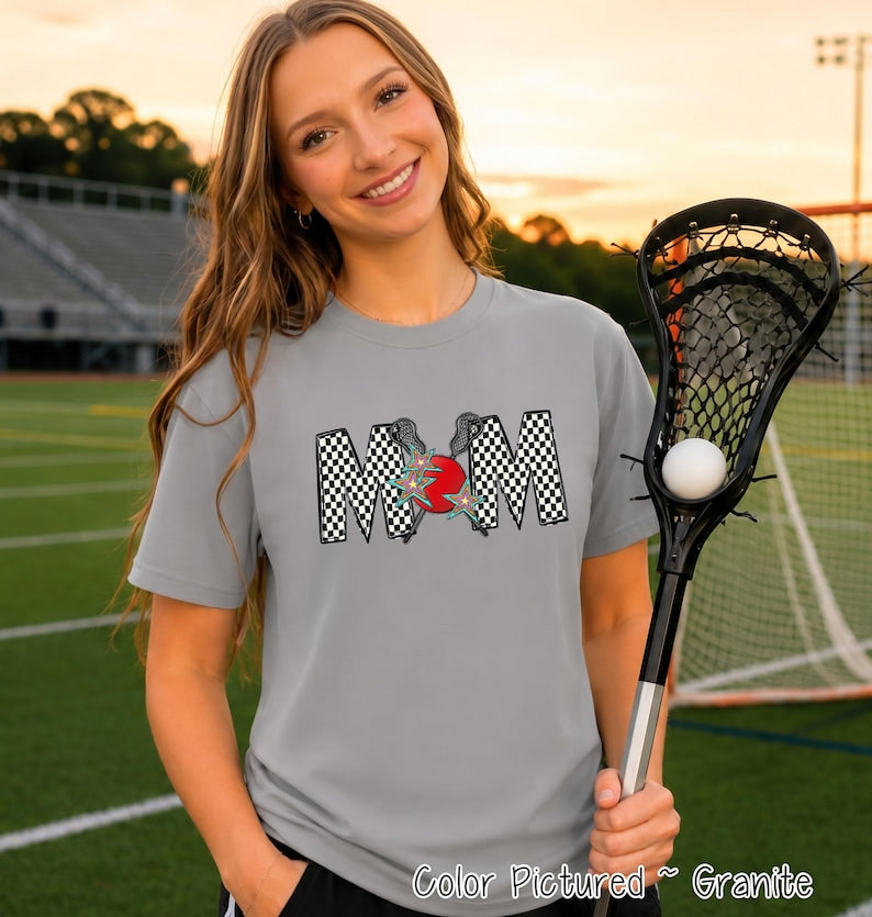 May include: A granite gray short-sleeve t-shirt featuring a graphic print of the word MOM in a black and white checkered pattern. The letter O is replaced by a red circle with two crossed lacrosse sticks and two teal stars layered on top.