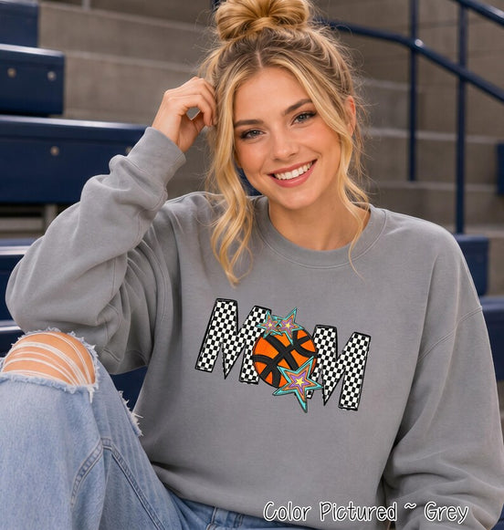 May include: A grey crewneck sweatshirt featuring the word MOM in a black and white checkered pattern. The letter O is replaced by an orange basketball graphic with black lines, accented by three multi-colored star shapes in blue, purple, and yellow.