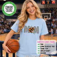 May include: A light blue short-sleeve t-shirt featuring the word MOM in a black and white checkered pattern, with an orange basketball replacing the letter O and two colorful stars. The shirt includes a Comfort Colors logo and text.