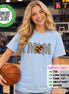 May include: A light blue short-sleeve t-shirt featuring the word MOM in a black and white checkered pattern, with an orange basketball replacing the letter O and two colorful stars. The shirt includes a Comfort Colors logo and text.