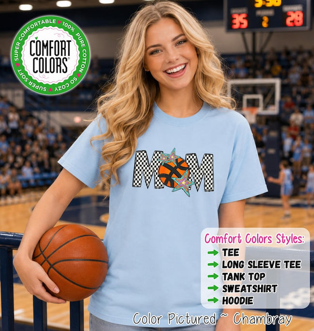 May include: A light blue short-sleeve t-shirt featuring the word MOM in a black and white checkered pattern, with an orange basketball replacing the letter O and two colorful stars. The shirt includes a Comfort Colors logo and text.