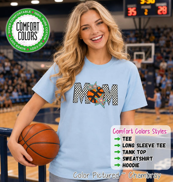 May include: A light blue short-sleeve t-shirt featuring the word MOM in a black and white checkered pattern, with an orange basketball replacing the letter O and two colorful stars. The shirt includes a Comfort Colors logo and text.