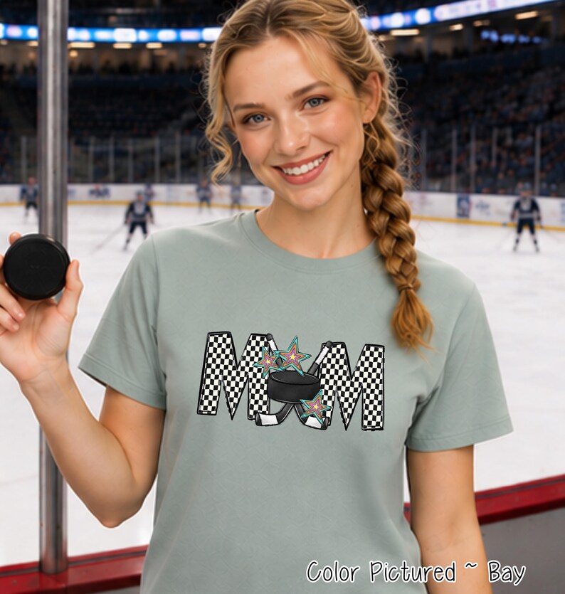 May include: A sage green short-sleeve t-shirt featuring a graphic print of the word MOM in a black and white checkered pattern. The letter O is replaced by a black hockey puck and two crossed hockey sticks, accented with colorful stars.