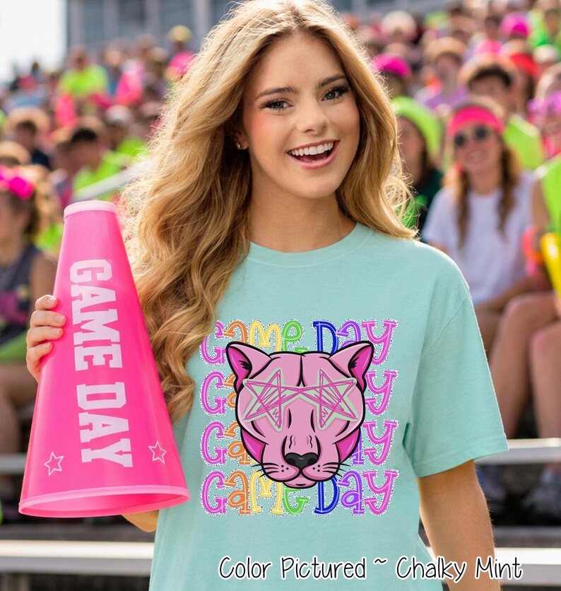 May include: A light mint green t-shirt featuring a graphic of a pink panther head wearing star-shaped glasses, surrounded by the repeating text "Game Day" in colorful, outlined letters. A bright pink megaphone with "GAME DAY" text is held.