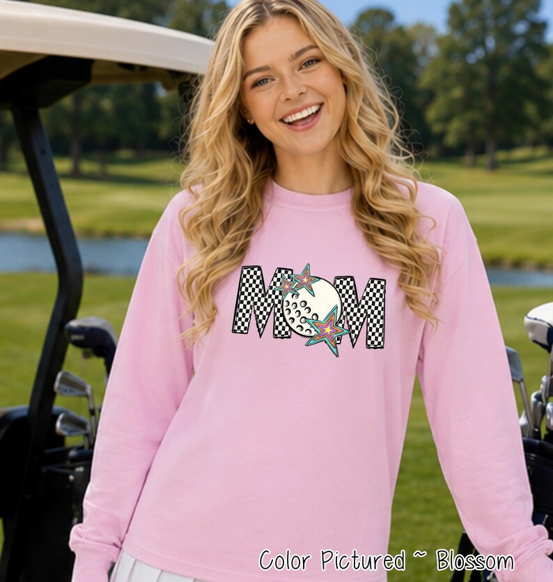May include: A light pink long-sleeve sweatshirt featuring a graphic on the chest that spells MOM in a black and white checkered pattern. The letter O is replaced by a white golf ball, and two colorful star shapes are layered over the letters.