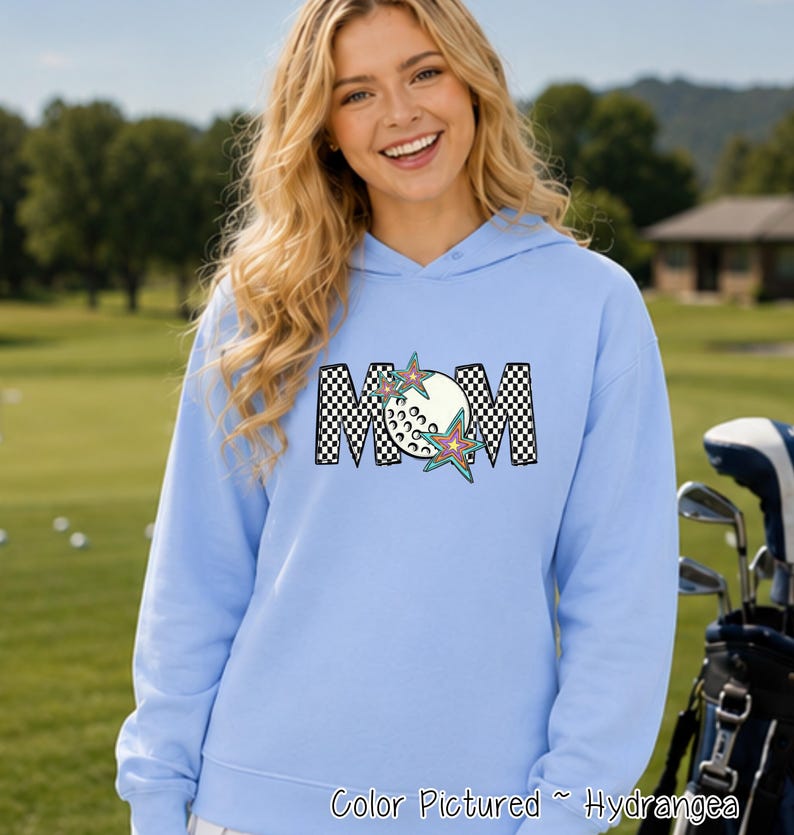 May include: A light blue hooded sweatshirt featuring the text MOM in a black and white checkered pattern. The letter O is replaced by a white golf ball graphic, and two colorful, multi-pointed stars are layered over the letters.