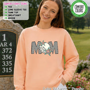 May include: A peach-colored crewneck sweatshirt featuring the word MOM in a black and white checkered pattern. The letter O is replaced by a white golf ball graphic, and two colorful, multi-pointed stars are layered over the letters.