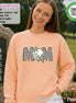 May include: A peach-colored crewneck sweatshirt featuring the word MOM in a black and white checkered pattern. The letter O is replaced by a white golf ball graphic, and two colorful, multi-pointed stars are layered over the letters.