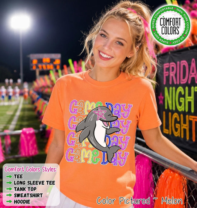 May include: An orange short-sleeve t-shirt featuring a graphic of a grey dolphin with a pink star over its eye. The text "GAME DAY" is repeated four times in colorful, outlined letters behind the dolphin. The shirt has a round neckline.