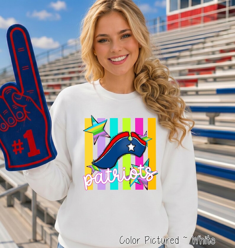 May include: A white long-sleeve sweatshirt featuring a graphic print of a blue, red, and white tricorn hat, colorful stars, and vertical stripes with the word patriots in white script. A blue foam finger with the number 1 is held up.